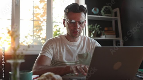 Handsome man works on his laptop at desk, with a cup of coffee and a pastry beside him. The evening sunlight fills the room, creating a serene and productive atmosphere in his home office workspace