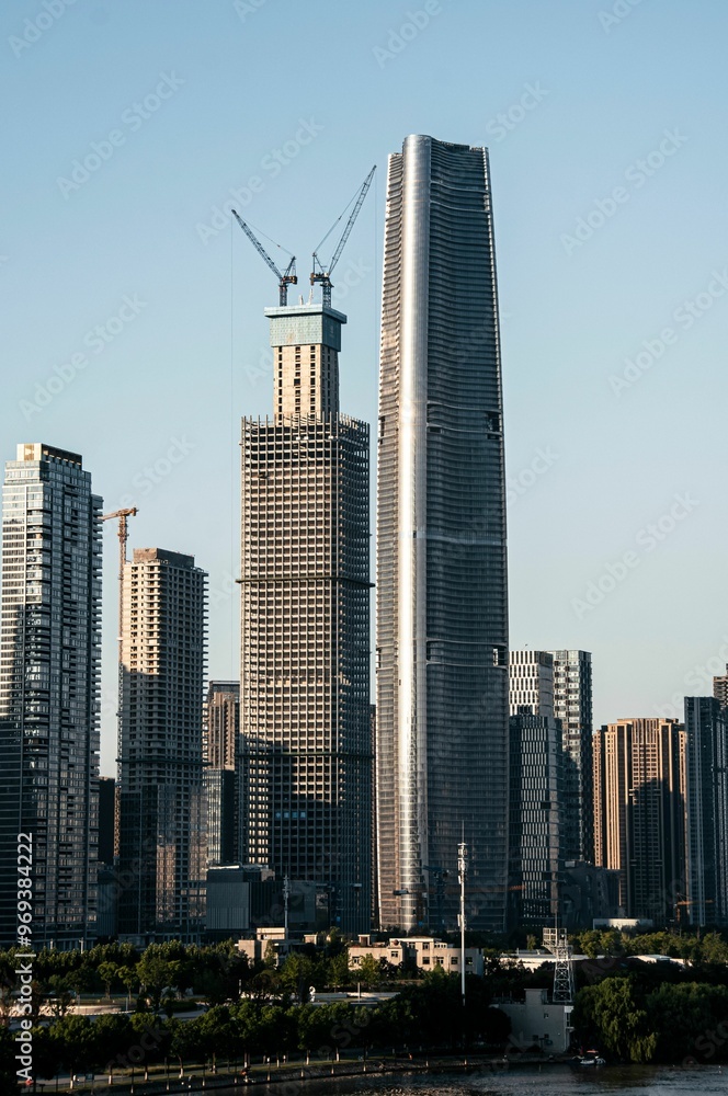 Fototapeta premium Skyscrapers in a city skyline with one under construction against a clear blue sky.