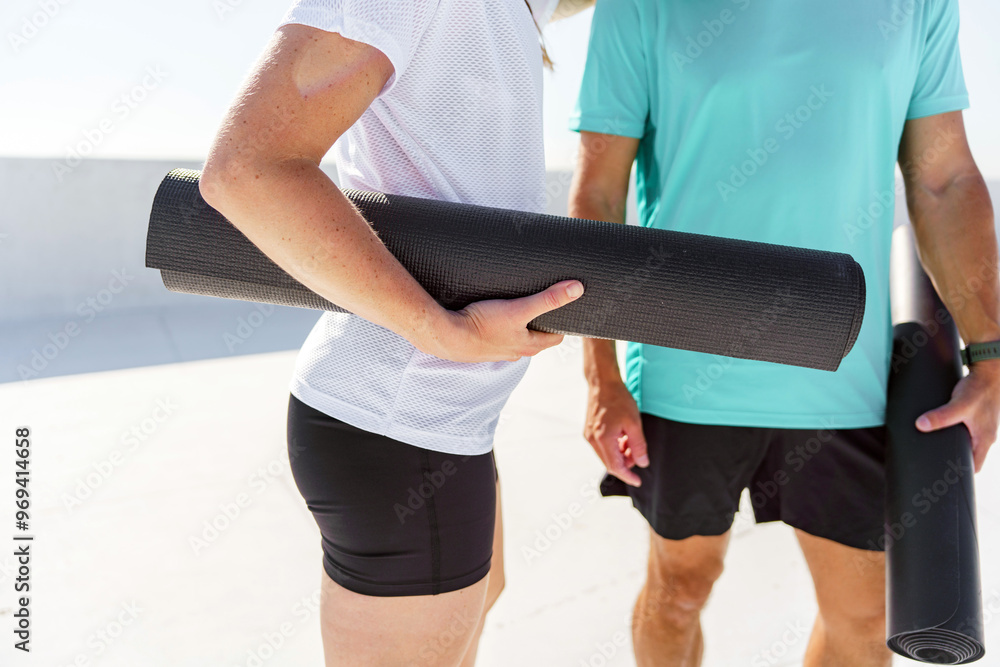 A close-up of two people holding rolled-up yoga mats, preparing for an outdoor yoga session under the sun. A close-up of two people holding rolled-up yoga mats, preparing for an outdoor yoga session under the sun.
