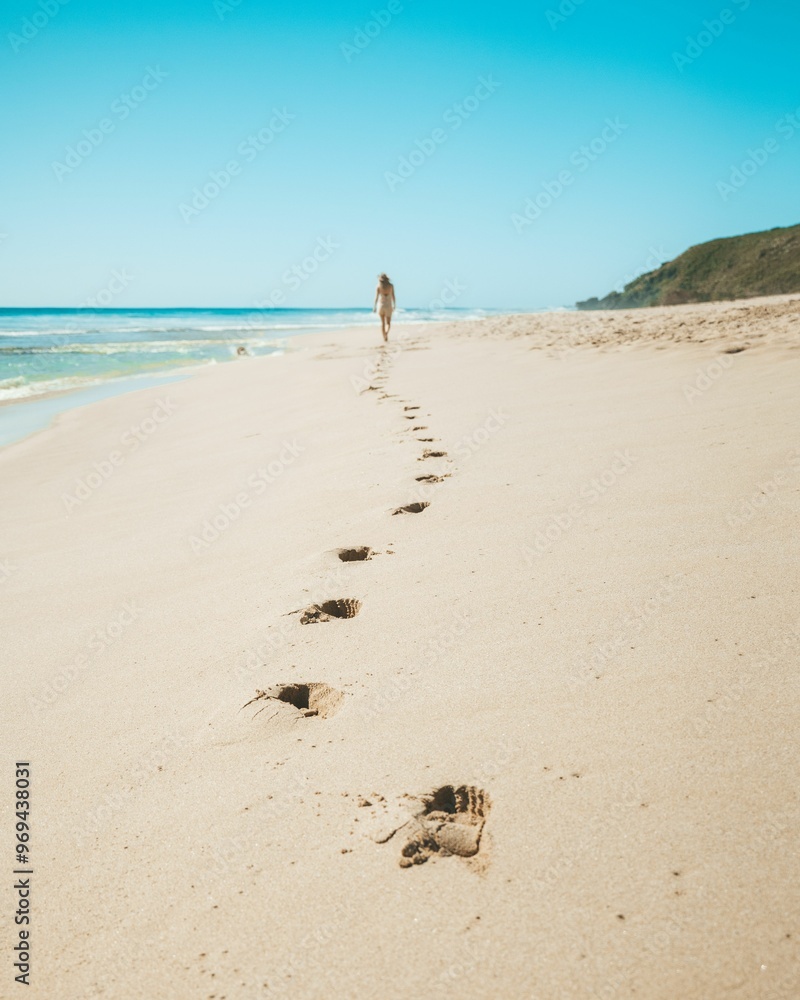 Woman walking along a sandy beach leaving footprints behind her, with a blue sky in the background Woman walking along a sandy beach leaving footprints behind her, with a blue sky in the background