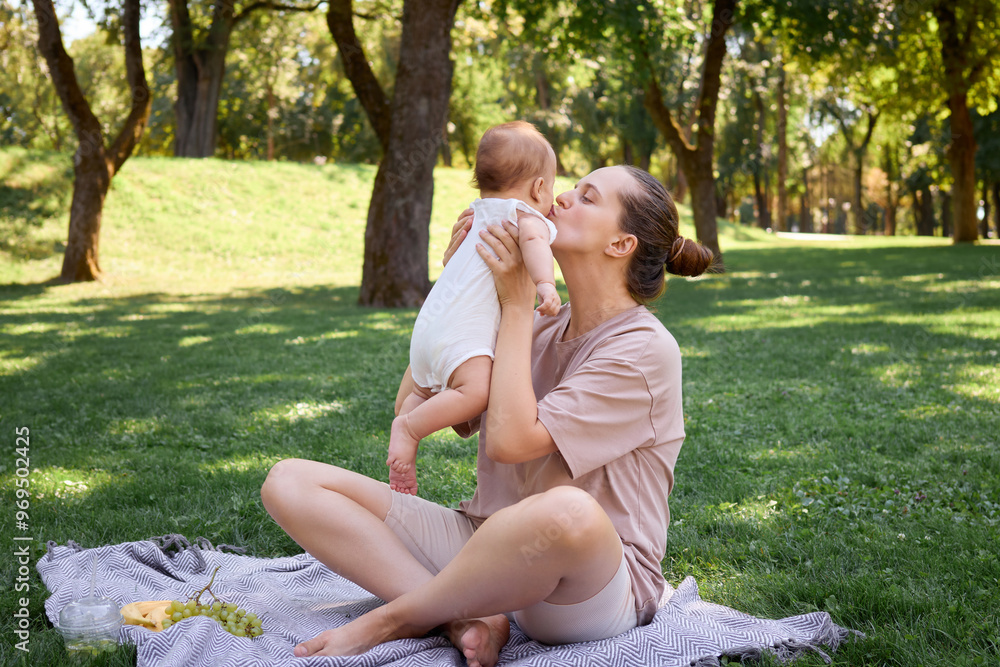 Fototapeta premium Loving mother kissing her infant child having fun picnic outdoors family bonding during vacation sitting on the grass enjoying healthy snacks and the joyful moments of happy day in park