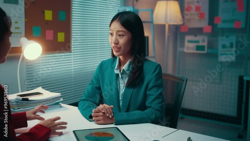 Two businesswomen are having a meeting in an office at night. They are discussing strategy and looking at some documents