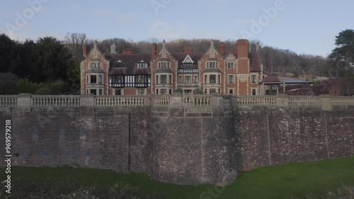 Aerial rising past brick wall to reveal Wood Norton Hall near Worcester in England with tall trees and light blue sky in the background
