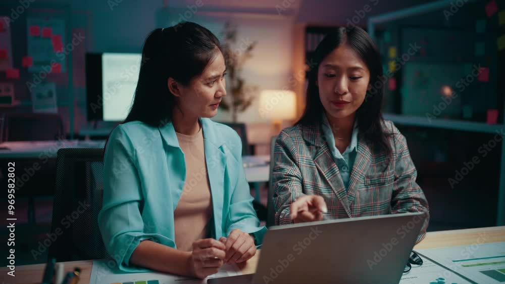 Two asian businesswomen are working late in the office. They are discussing something on a laptop computer and appear to be enjoying their work Two asian businesswomen are working late in the office. They are discussing something on a laptop computer and appear to be enjoying their work