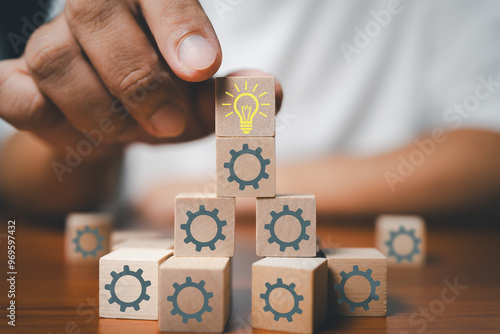 Hand placing a wooden block with a lightbulb icon on top of a stack of blocks with gear icons