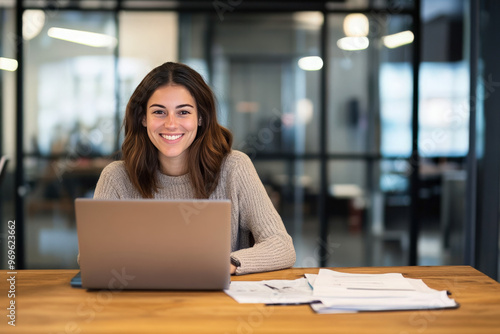 A young woman smiles confidently while working on her laptop at a desk in a modern office. Free Download A young woman smiles confidently while working on her laptop at a desk in a modern office. - GFXLoad