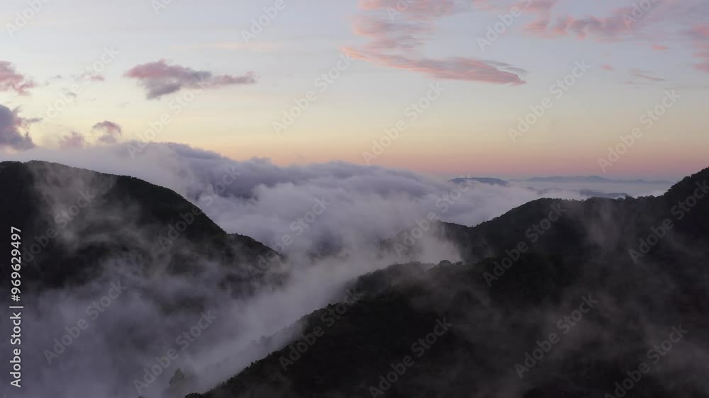 Aerial descending through wispy clouds towards mountain forest landscape and with pink and blue soft dawn sunrise in the background Aerial descending through wispy clouds towards mountain forest landscape and with pink and blue soft dawn sunrise in the background