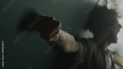 Rack focus shot of young man screaming and pounding dark grey wall with edge of palm clenched in fist in despair
