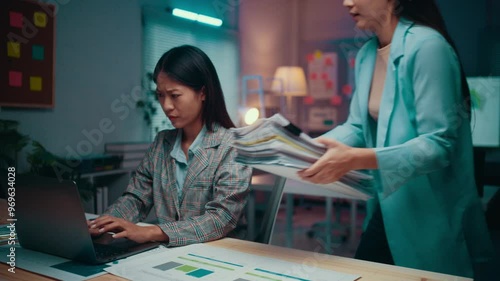 Manager is bringing a stack of paper to her employee working overtime in a dark office. The employee is overwhelmed with work