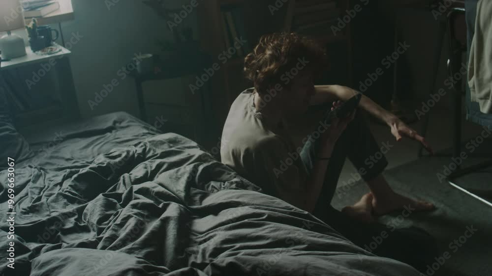 Young man having emotional phone conversation and throwing it on messy bed, then putting head on arms while sitting with bent legs on carpet on bedroom floor Young man having emotional phone conversation and throwing it on messy bed, then putting head on arms while sitting with bent legs on carpet on bedroom floor