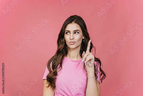 A woman in a pink top stands against a pink background, raising her index finger and looking upward, suggesting an idea or inspiration.