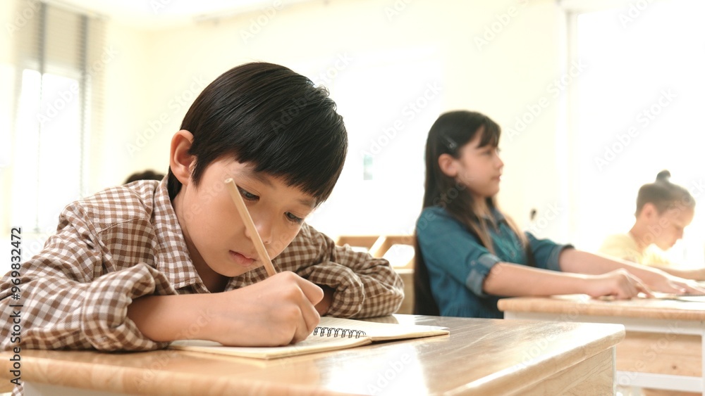 Close up of skilled asian student writing or taking a note during test. Elementary student working on paper while multicultural doing classwork or homework at classroom. Education concept. Pedagogy. Close up of skilled asian student writing or taking a note during test. Elementary student working on paper while multicultural doing classwork or homework at classroom. Education concept. Pedagogy.