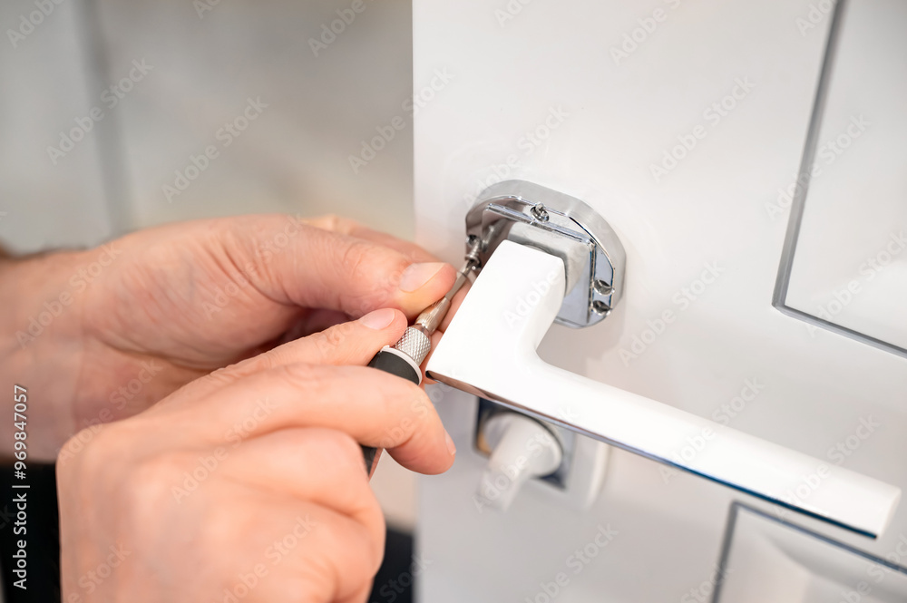 A locksmith adjusts the door handle of a modern white interior door. A screwdriver and a man's hands in close-up A locksmith adjusts the door handle of a modern white interior door. A screwdriver and a man's hands in close-up