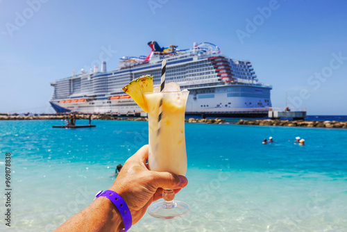 Close-up view of hand holding glass of alcoholic cocktail on Caribbean beach with cruise ship in background. Curacao.
