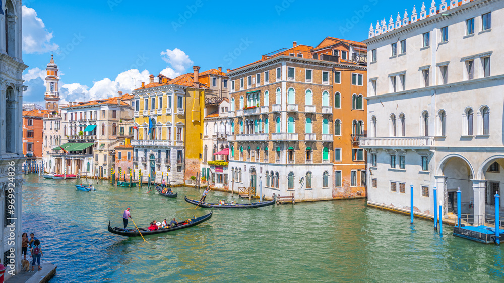 Fototapeta premium Brightly colored buildings line the Canal Grande as gondolas glide through the water under a clear blue sky. Tourists enjoy the vibrant atmosphere of Venice during a perfect day.
