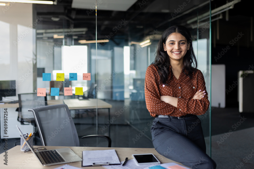 © Liubomir - Portrait of young beautiful Latin American business woman, company employee smiling and looking at camera with crossed arms, businesswoman satisfied with work inside office using laptop.