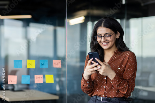 Young business woman using phone inside office, company worker smiling typing text message and browsing social networks, hispanic woman satisfied with work standing near window.