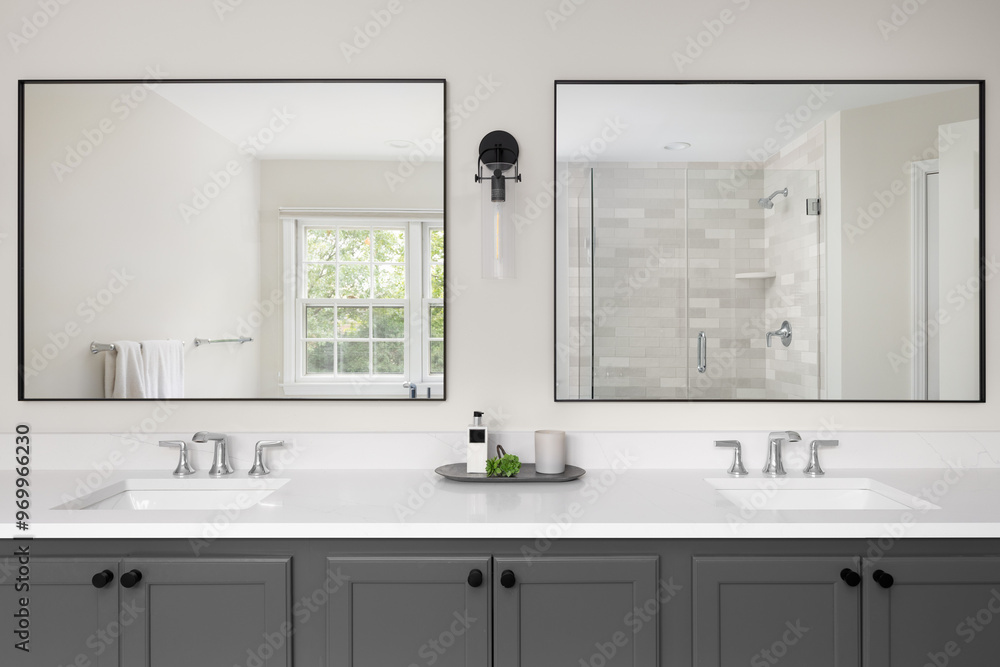 A bathroom with a grey vanity cabinet, white marble countertop with two sinks, and a view through the large mirrors of the tiled shower and window. A bathroom with a grey vanity cabinet, white marble countertop with two sinks, and a view through the large mirrors of the tiled shower and window.