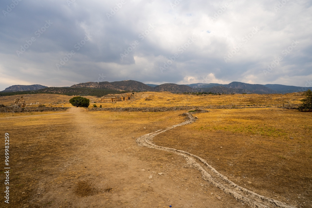 Hierapolis in Pamukkale: A desolate path winds through the arid landscape, leading towards ancient ruins nestled beneath a dramatic, cloud-filled sky.