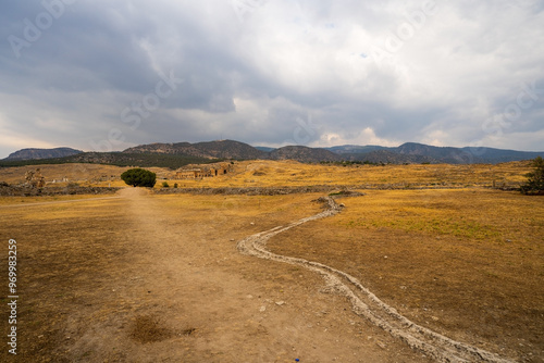 Hierapolis in Pamukkale: A desolate path winds through the arid landscape, leading towards ancient ruins nestled beneath a dramatic, cloud-filled sky.