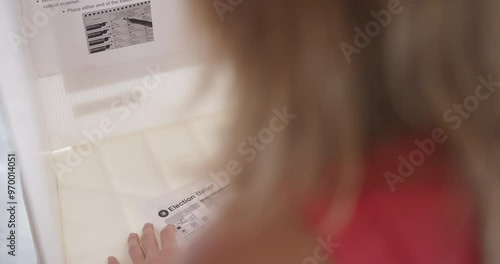 Close, over the shoulder view of young woman voter filling in a ballot at a voting booth in the USA. Hand held camera on stabilized gimbal. Candidate names obscured. 4K DCI