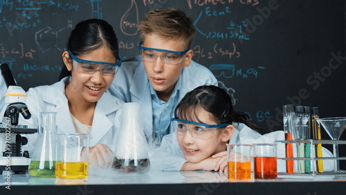 Girl looking at beaker while doing experiment at blackboard with chemistry theory in STEM class. teacher and group of highschool student with mixed races doing science activity together. Edification.