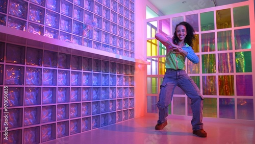 Happy hispanic girl in lively mood moving along music in neon light while wearing colorful cloth at night club. Street dancer making energetic footstep or movement while looking at camera. Regalement.