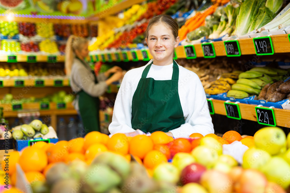 Obraz premium Portrait of smiling young girl wearing apron standing in fruit and vegetable store. First job concept..