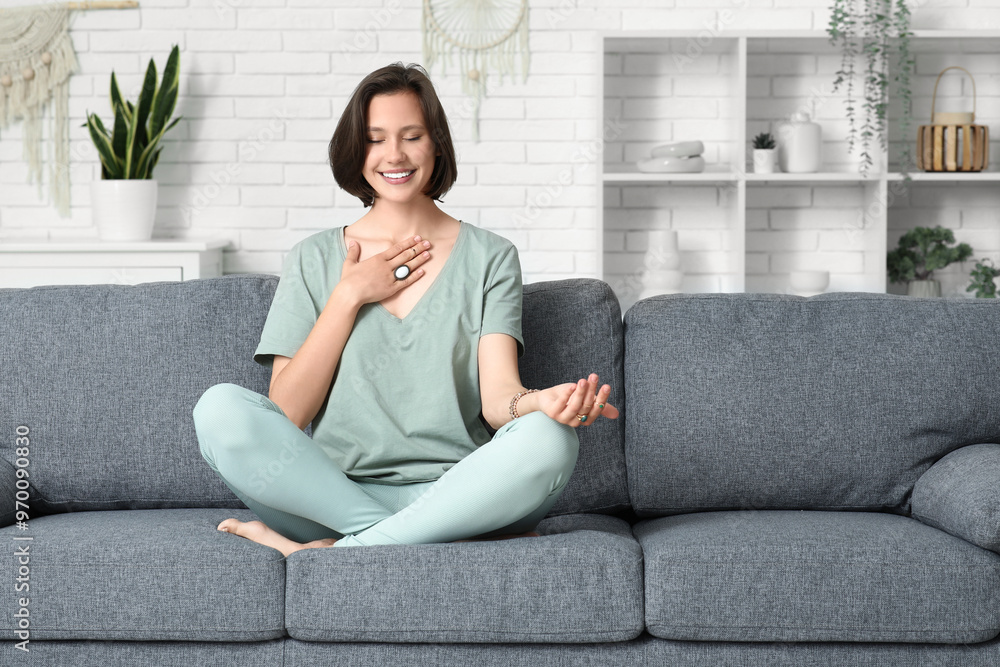Fototapeta premium Young woman meditating while sitting on sofa at home