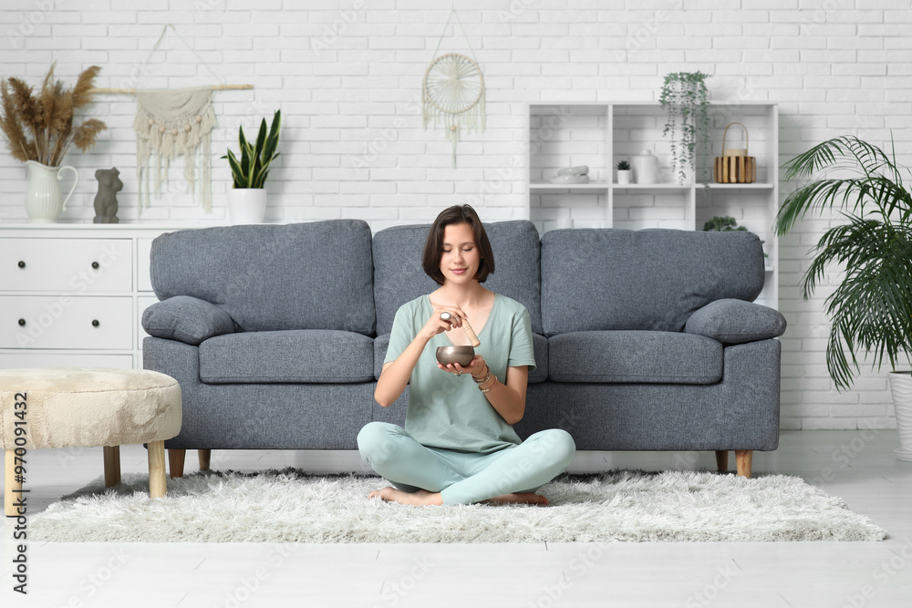 Obraz premium Young woman with Tibetan singing bowl meditating while sitting on floor at home