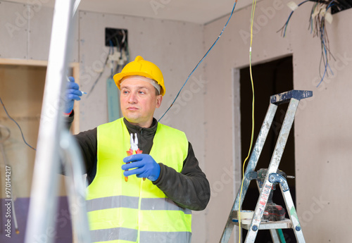 Repair man in yellow hardhat and vest working on electric wires during reconditioning works in apartment. photo