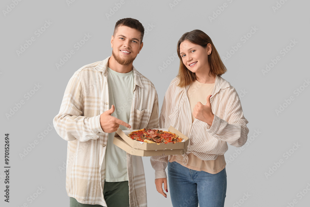 Beautiful young happy couple pointing at cardboard box of tasty pizza and showing thumb-up gesture on grey background Beautiful young happy couple pointing at cardboard box of tasty pizza and showing thumb-up gesture on grey background