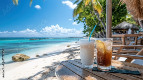 Two refreshing beverages, one with a straw, are placed on a wooden table by the beach, with a stunning blue ocean, lush palm trees, and a clear sky creating a perfect tropical setting.