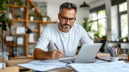 A man wearing glasses, focusing deeply on his work, writes on paper while using a laptop in a home office surrounded by plants and natural light.