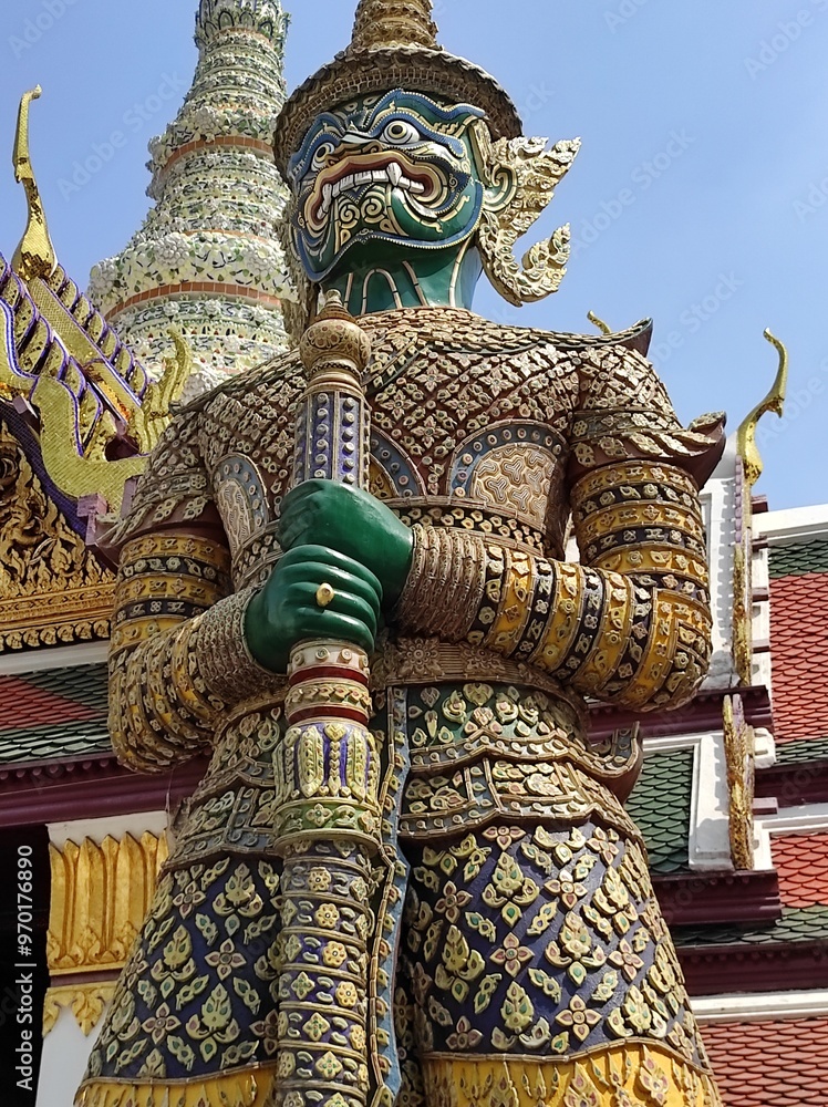 An imposing and vividly decorated giant guardian statue at Wat Phra Kaew, featuring a detailed mosaic of colorful patterns, standing guard with a backdrop of golden spires and traditional Thai archite An imposing and vividly decorated giant guardian statue at Wat Phra Kaew, featuring a detailed mosaic of colorful patterns, standing guard with a backdrop of golden spires and traditional Thai archite
