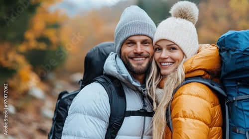 A smiling and happy couple dressed warmly in puffer jackets and woolen hats are hiking through an autumn landscape with their backpacks, enjoying nature and each other’s company.