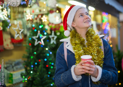 Happy teenage girl wearing traditional Santa hat and gold tinsel around her neck walking in colorful Christmas street market, holding paper cup of warm drink photo