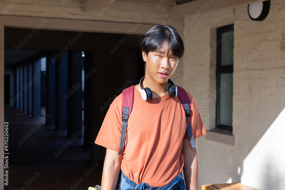 Obraz premium Asian teenage boy with headphones and backpack near school building, looking ahead