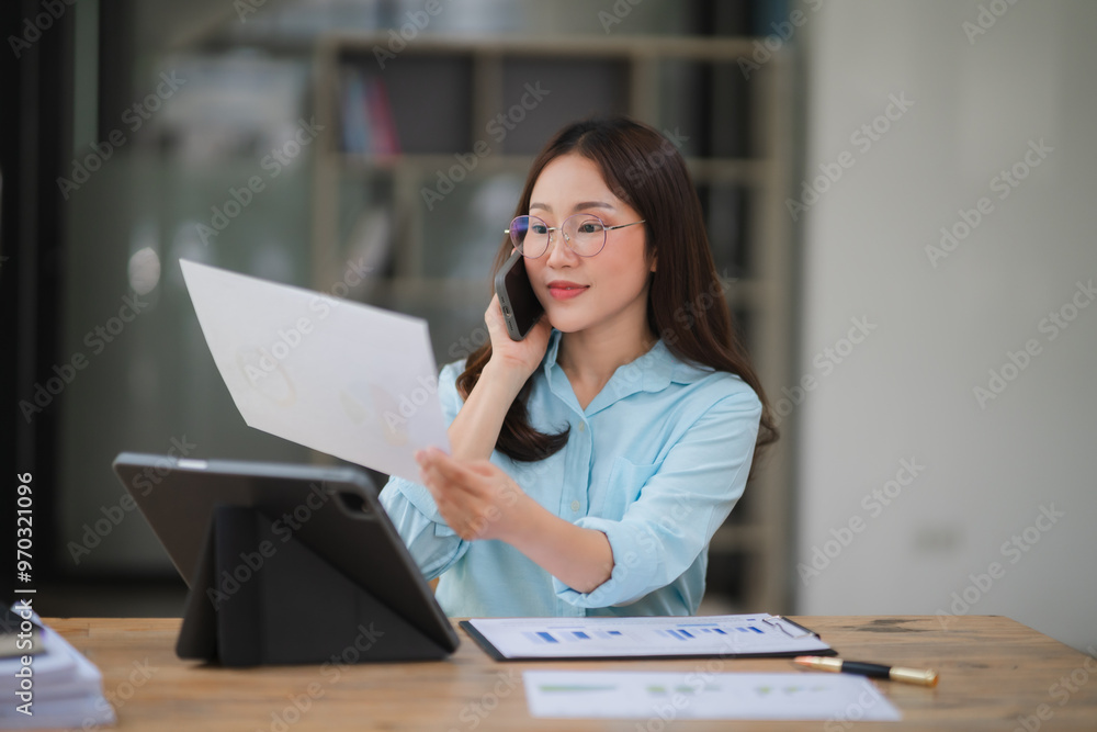 Businesswoman on Phone Call: A focused young businesswoman in a blue shirt sits at a desk in a modern office, reviewing documents while on a phone call. She exudes professionalism and efficiency, show Businesswoman on Phone Call: A focused young businesswoman in a blue shirt sits at a desk in a modern office, reviewing documents while on a phone call. She exudes professionalism and efficiency, show