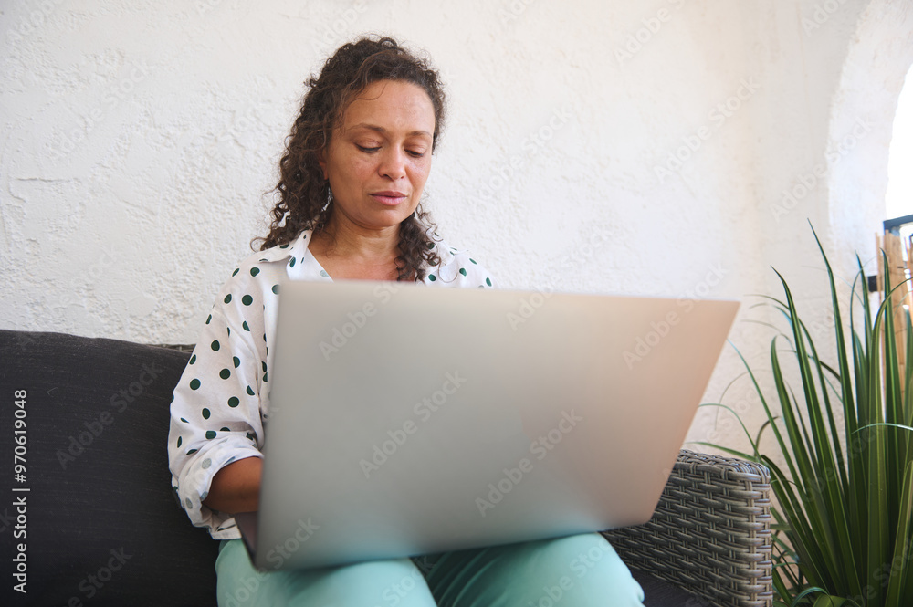 Woman working on a laptop in a cozy home setting, focusing on her tasks, wearing a polka dot shirt. Relaxed and productive atmosphere with natural light.
