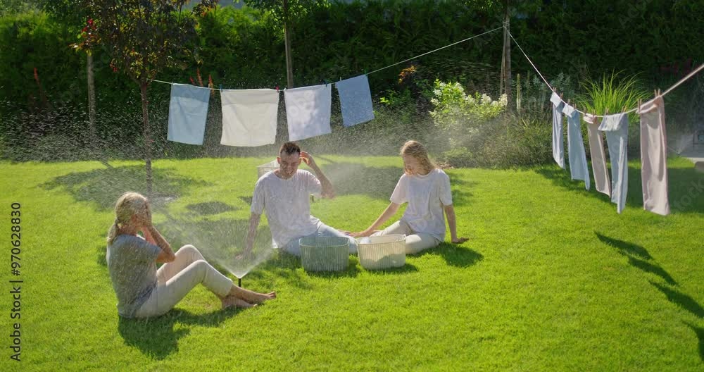 A mother with her two children sits on the lawn as the automatic sprinkler turns on, creating a fun moment in the hot summer day. They are laughing and playing together A mother with her two children sits on the lawn as the automatic sprinkler turns on, creating a fun moment in the hot summer day. They are laughing and playing together