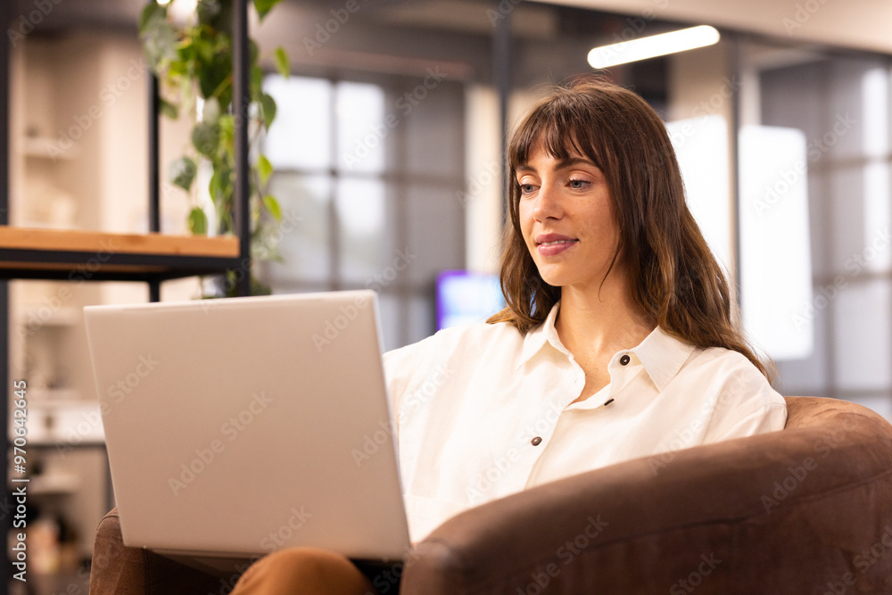 Working on laptop, woman sitting on chair in modern office environment Working on laptop, woman sitting on chair in modern office environment