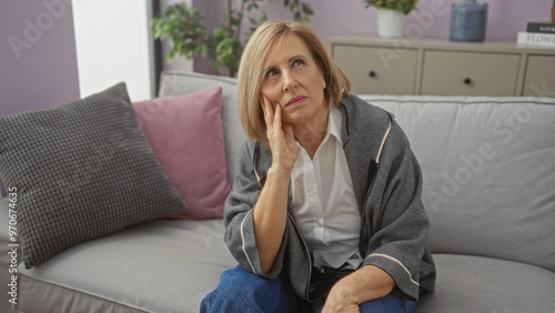 Mature blonde woman sitting pensively on a couch in a cozy living room, adorned with cushions and plants in the background.