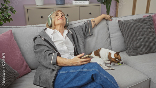 A mature blonde woman wearing headphones relaxes on a couch with her small dog in a cozy living room, holding a smartphone and enjoying a tranquil indoor moment. photo