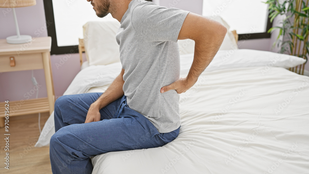 A young bearded man in casual attire sits on a bed with a pained expression, holding his lower back in a well-lit bedroom. A young bearded man in casual attire sits on a bed with a pained expression, holding his lower back in a well-lit bedroom.