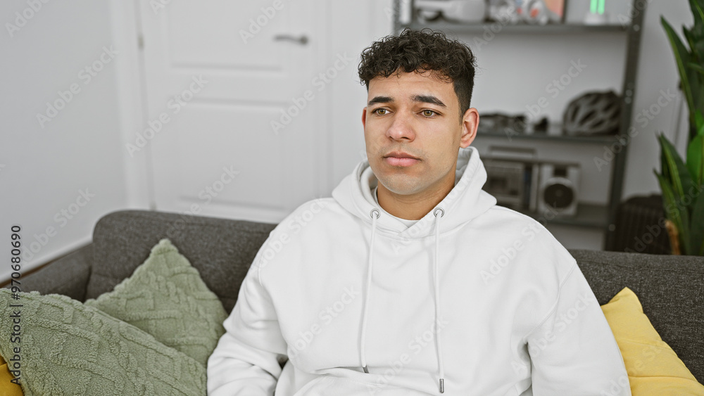 Young man with curly hair in a white hoodie sitting casually on a couch in a modern living room Young man with curly hair in a white hoodie sitting casually on a couch in a modern living room