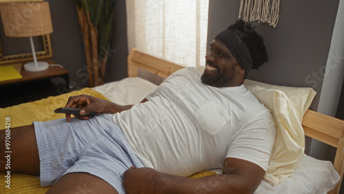 A handsome african american man with a beard lies on a bed in his bedroom, smiling, and holding a remote control, enjoying a relaxed moment indoors.