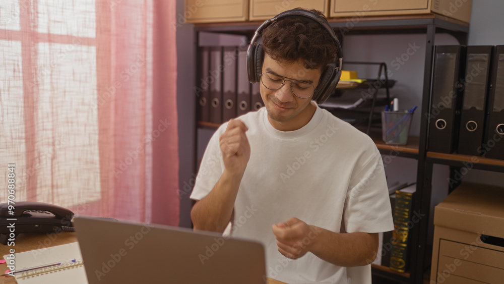 Young hispanic man mimics playing the drums while wearing headphones in an office environment, surrounded by folders and boxes. Young hispanic man mimics playing the drums while wearing headphones in an office environment, surrounded by folders and boxes.