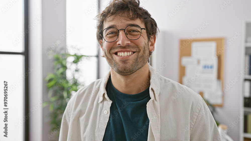 A smiling bearded man wearing glasses is standing in a bright modern office. A smiling bearded man wearing glasses is standing in a bright modern office.