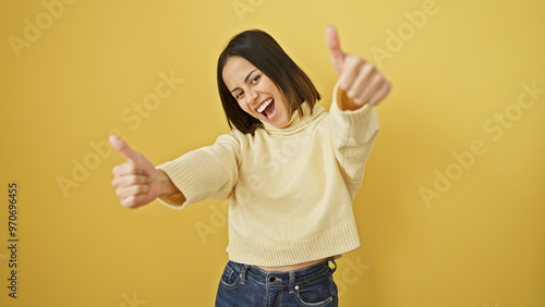 A cheerful young hispanic woman gives two thumbs up against a vibrant yellow wall, embodying positivity and approval.